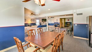 A hotel room with a wooden dining table, four chairs, a ceiling fan, and two beds with blue bedding. - Relaxing Surfside Bunkhouse - Walk to the Beach! (Freeport)