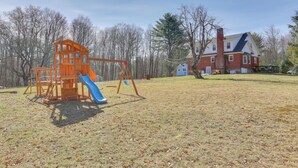 A playground with a slide and swings, a brick house with a chimney, and leaf-covered ground. - Cozy Catskills Tiny Home ~ 3 Mi to Bethel Woods! (Swan Lake)