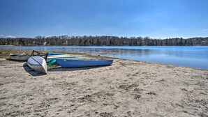 A sandy beach with several boats, a calm lake, and a forested shoreline under a clear sky. - Spacious Tobyhanna Family Home: Pool/beach Access! (Tobyhanna)