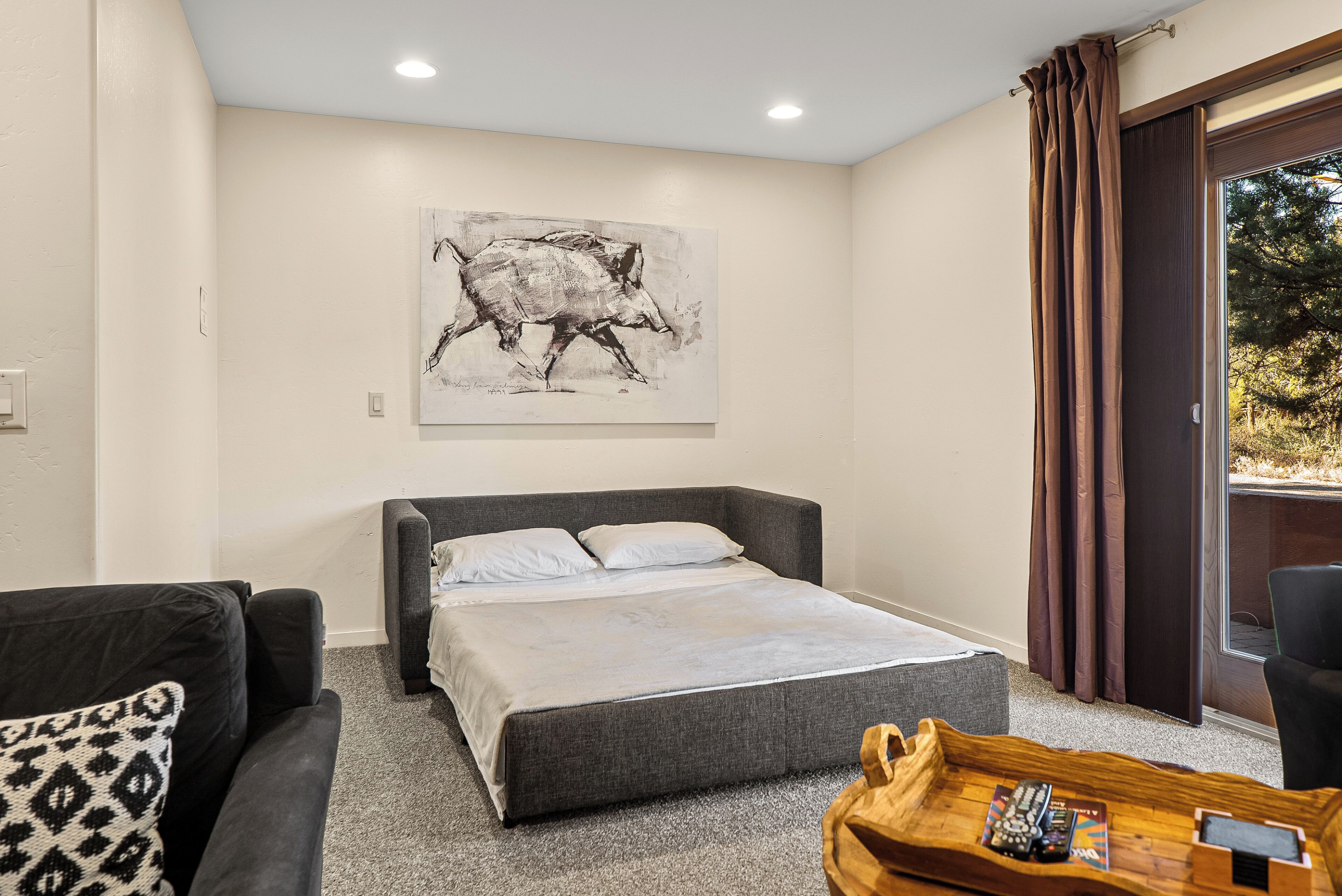 A bedroom with a bed, a sofa, a wooden coffee table with remote controls, and a large window with curtains.