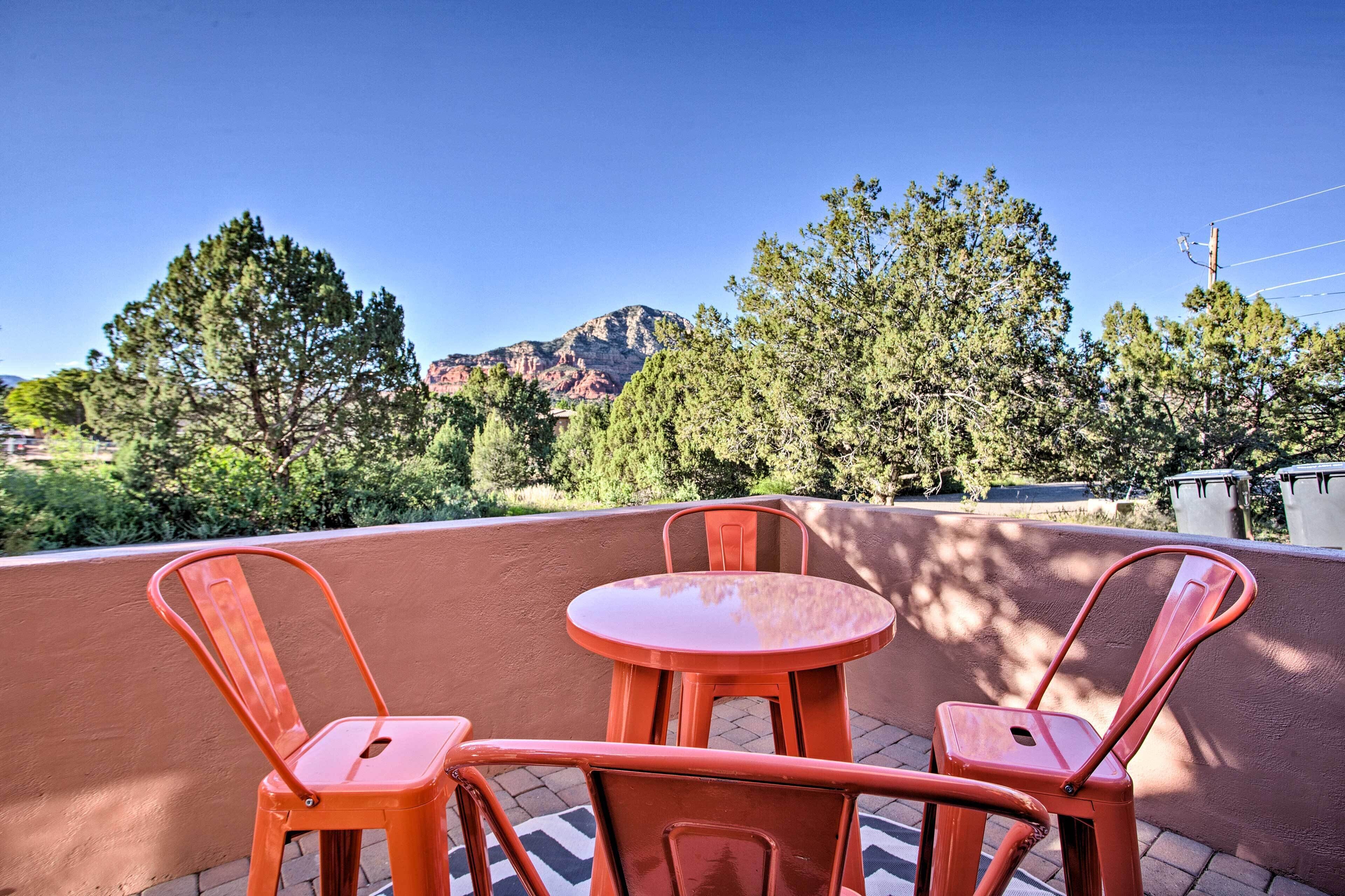 A patio with two red metal chairs and a small round table, overlooking a mountainous landscape with trees and a clear sky.