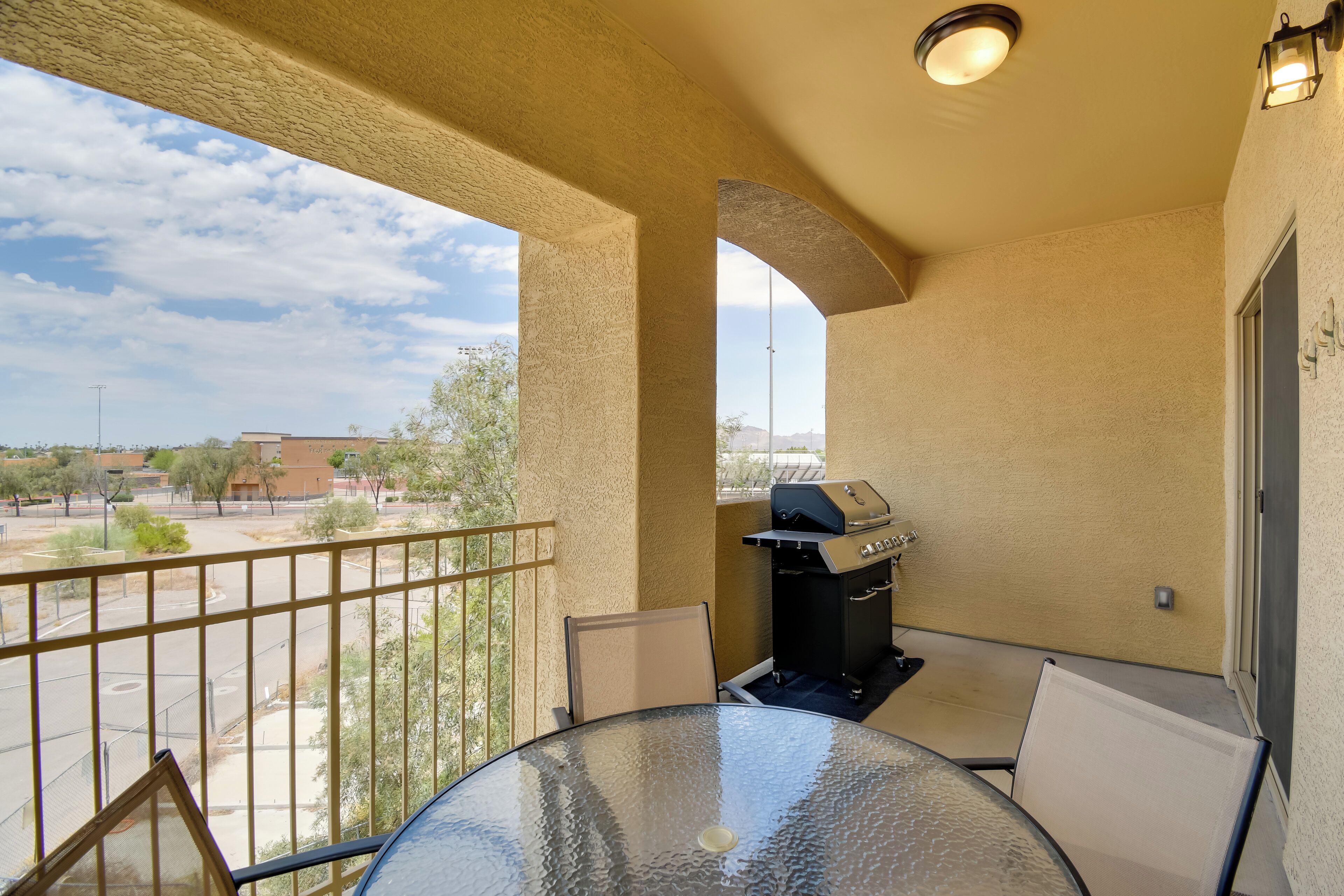 A balcony with a glass table and chairs, a grill, and a view of the street and buildings.