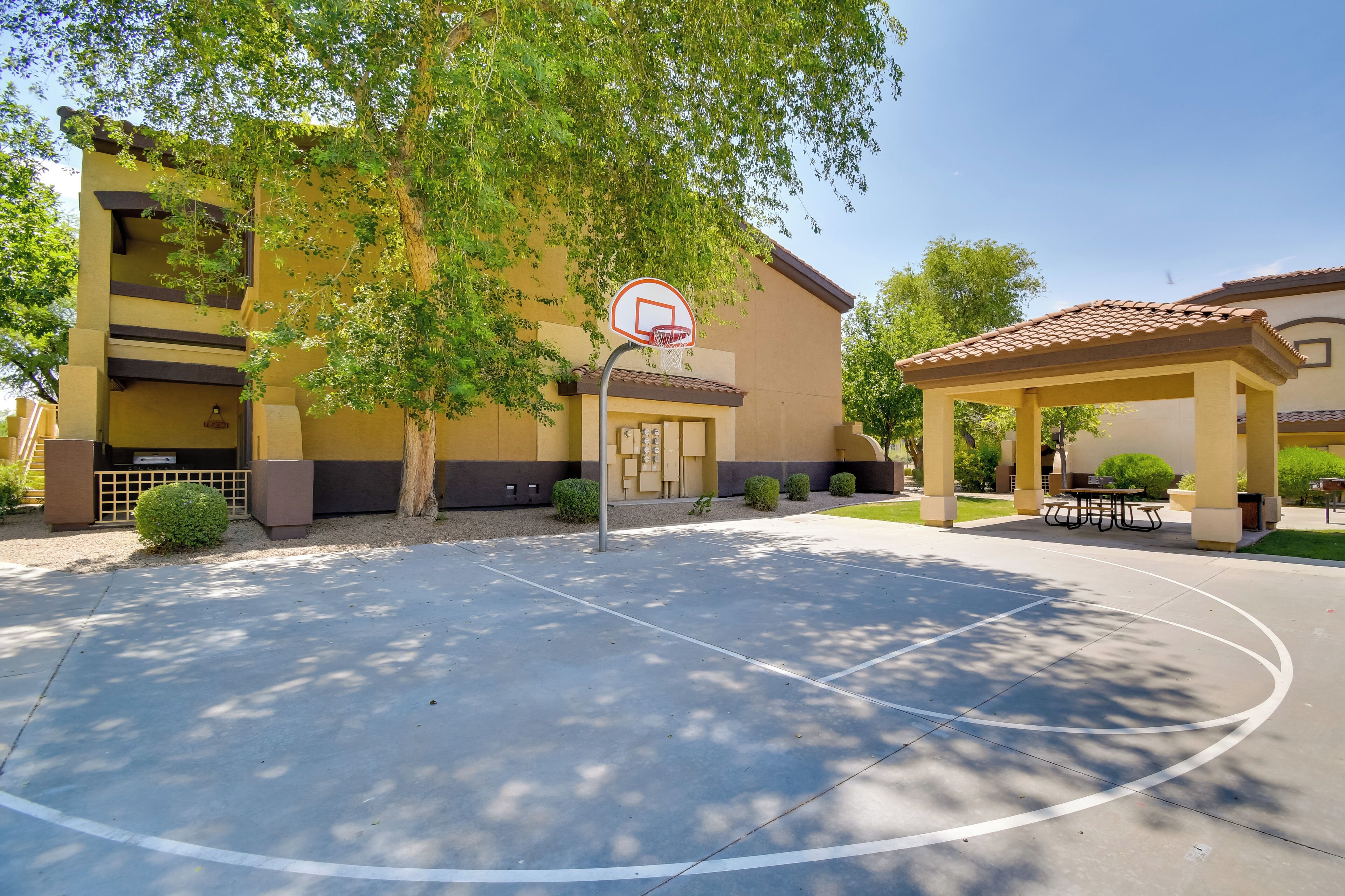 Ein Basketballplatz im Freien mit Korb, umgeben von Bäumen und einem Gebäude mit gelber Fassade.