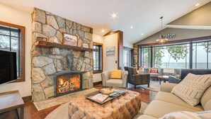 A cozy living room with a stone fireplace, comfortable seating, and a large window overlooking a body of water. - 1 Mi to Dtwn! Lakefront Earl Young Charlevoix Home (Charlevoix)