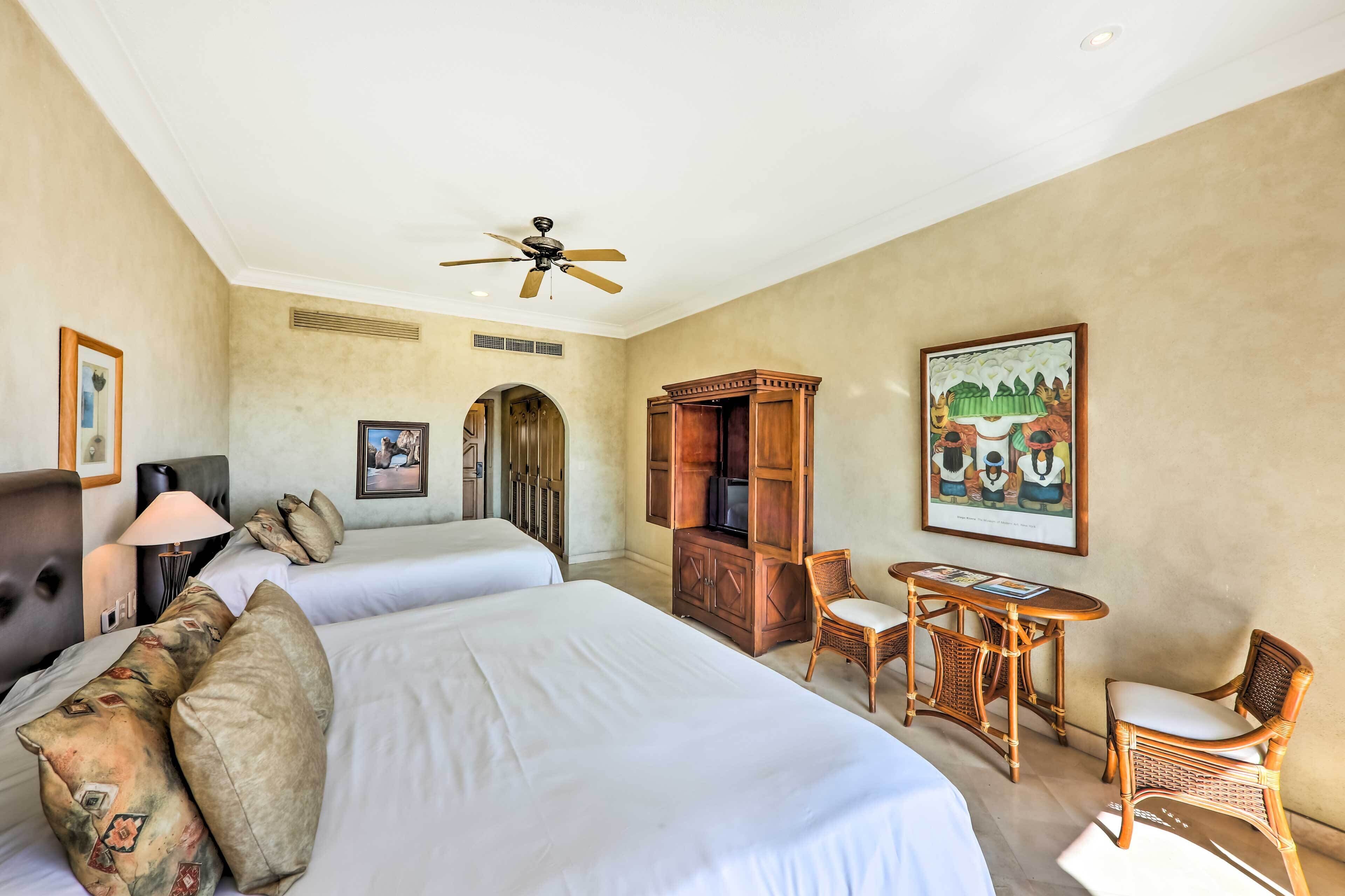 Habitación de hotel con dos camas, ventilador de techo, televisor, mesa de comedor con sillas y obras de arte en las paredes.