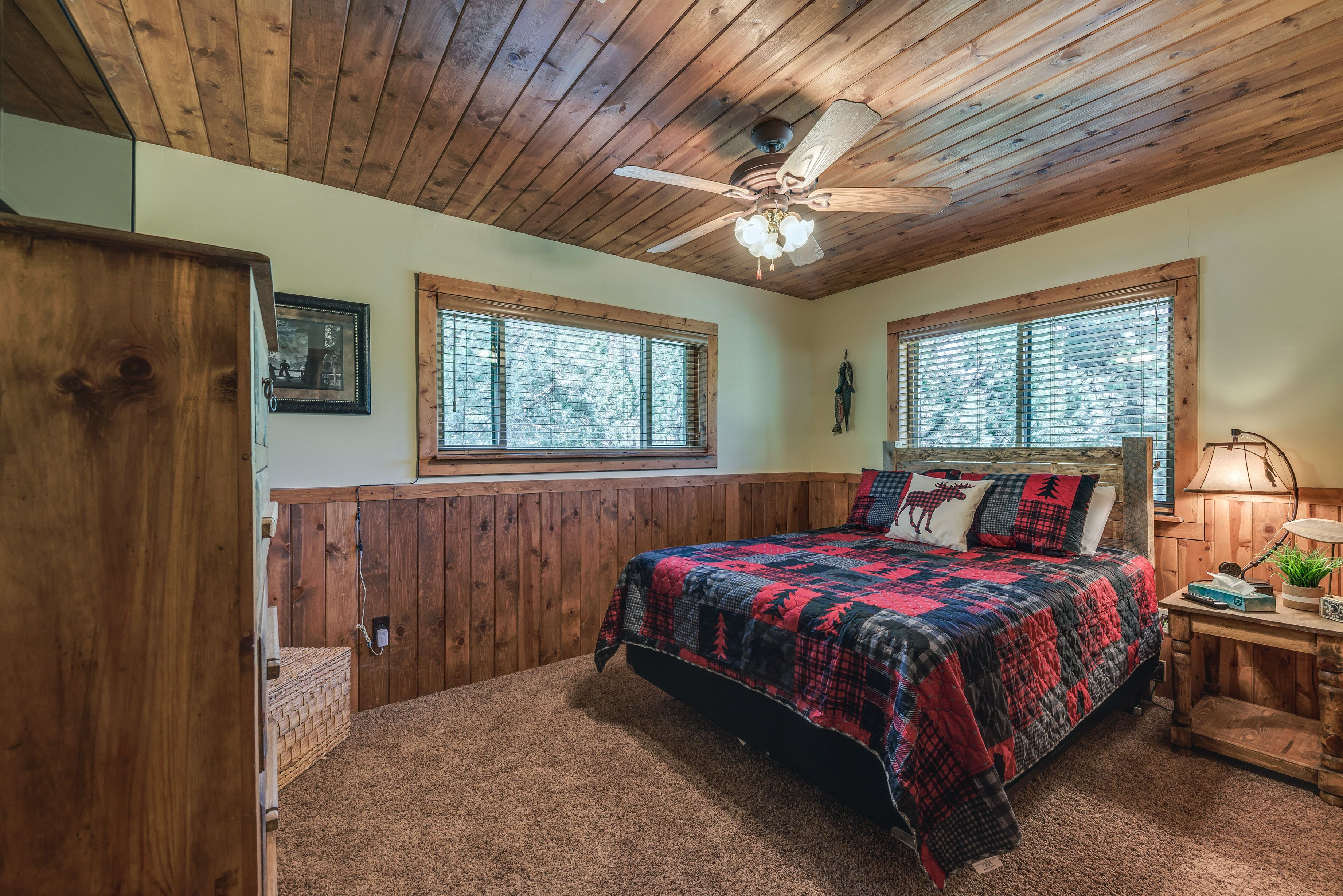 A bedroom with a wooden ceiling, a bed with a red and black quilt, two windows, a ceiling fan, and a nightstand with a lamp.