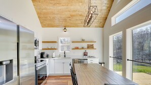 A modern kitchen with white cabinetry, a wooden dining table, and a view of the outdoors through large windows. - Modern Appalachian Vacation Rental w/ River Access (Pennington Gap)