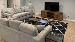 Ein Wohnzimmer mit grauem Sofa, einem Couchtisch, einem Fernseher auf einem Holzständer und einem gemusterten Teppich.