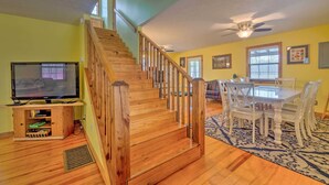 Eine Holztreppe führt zu einer zweiten Etage, einem Essbereich mit weißen Stühlen, einem Deckenventilator und einem Fernseher auf einem Holzständer.