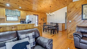 A cozy cabin interior with a wooden ceiling, a dining area, a kitchen with a refrigerator, and a leather sofa with a bear pillow. - Bryson City Cabin w/ Hot Tub, Views & Pool Table! (Bryson City)