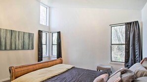 Un dormitorio con una cama grande, una mesita y dos ventanas con cortinas.