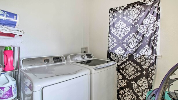 Un área de lavandería con dos lavadoras Samsung, un estante con productos de limpieza y una cortina estampada.