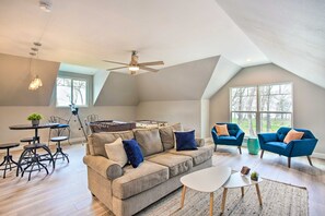 Un loft espacioso con techo alto, un ventanal grande, un sofá, dos sillones, una mesa de centro y una zona de comedor con mesa y sillas.