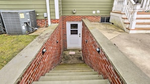 Um conjunto de degraus de concreto leva a uma porta branca, ladeada por paredes de tijolos vermelhos, com aparelhos de ar condicionado e um pequeno degrau de tijolos visíveis em primeiro plano.