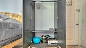 Una habitación compacta con una cama, un pequeño kitchenette con ollas, un tazón azul y una hornilla.