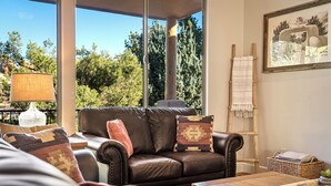 A living room with a leather sofa, a wooden ladder, and a large window with a view of trees. - Luxury Sedona Living: Remodeled w/ Red Rock Views! (Sedona)