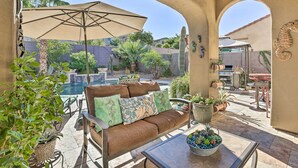 A patio area with a sofa, table, and umbrella, overlooking a pool and garden. - Luxe Goodyear Home w/ Serene Backyard & Game Room (Goodyear)