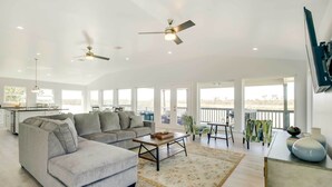 A spacious living room with a grey sectional sofa, a coffee table, and a large window offering a view of the outdoors. - Spacious San Jacinto River Home w/ Waterfront Deck (Houston)