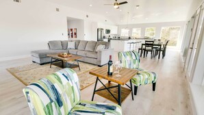A spacious living area with a grey sectional sofa, a wooden coffee table, and striped armchairs. A kitchen with white cabinets and a dining area with a wooden table and chairs are visible in the background. - Spacious San Jacinto River Home w/ Waterfront Deck (Houston)