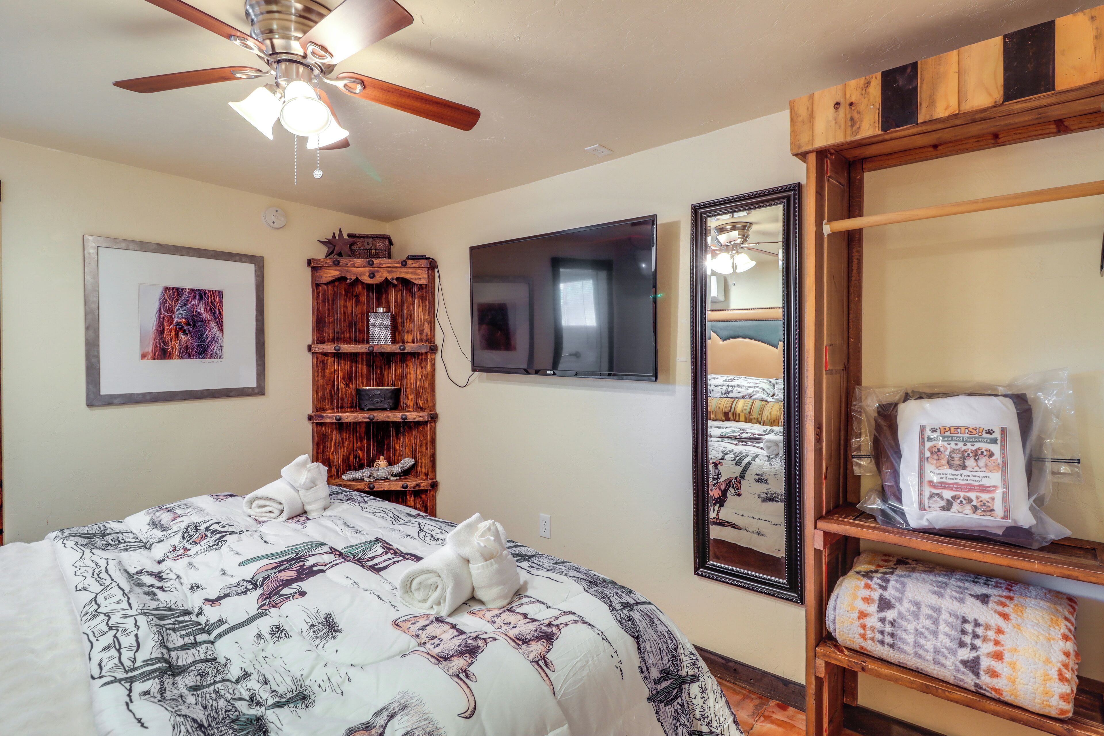 A hotel room with a bed, a ceiling fan, a TV, a wooden shelf with towels, a framed picture, and a mirror.