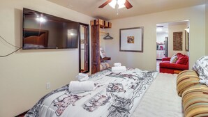 Un dormitorio con cama, ventilador de techo, espejo, una silla roja y un pasillo que lleva a una cocina.