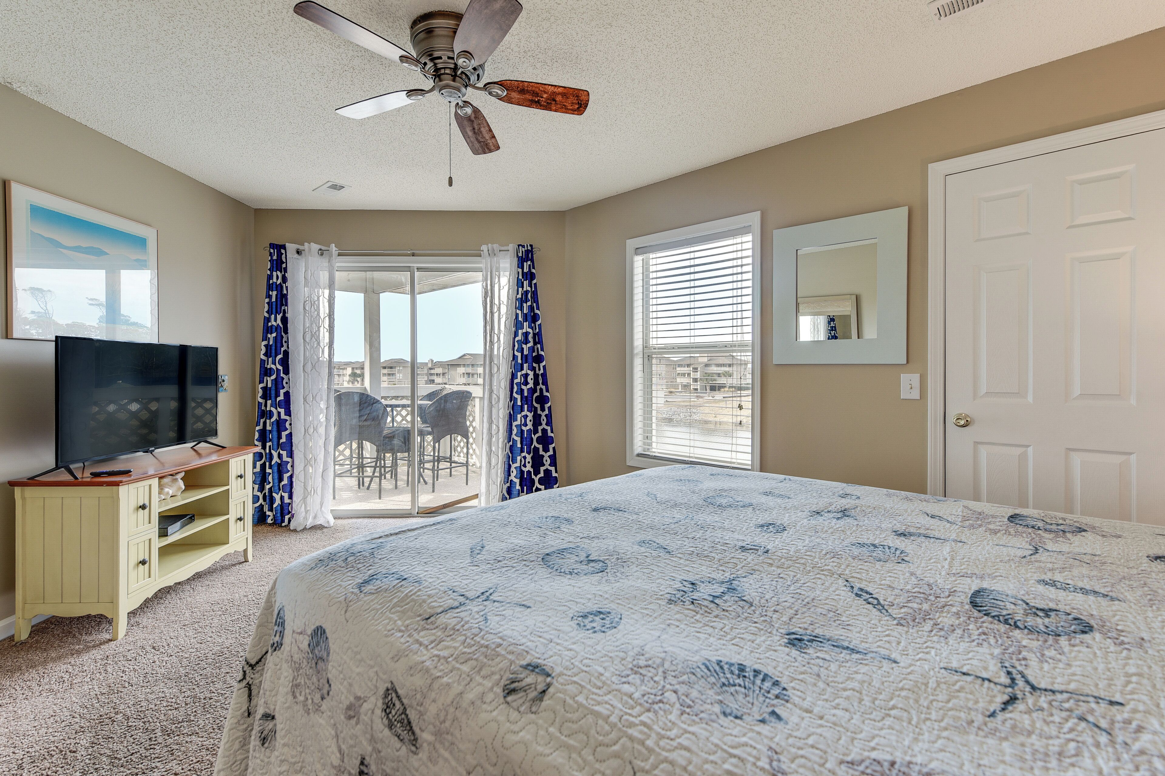 A bedroom with a large bed, a ceiling fan, a TV on a wooden cabinet, and a view through a glass door.