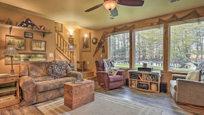 A cozy living room with a wooden floor, a leather sofa, a patterned armchair, and a wooden coffee table. There is a staircase with a wooden railing, a large window with a view of trees, and a ceiling fan. - Secluded Birnamwood Cottage w/ Deck & River Views! (Birnamwood)