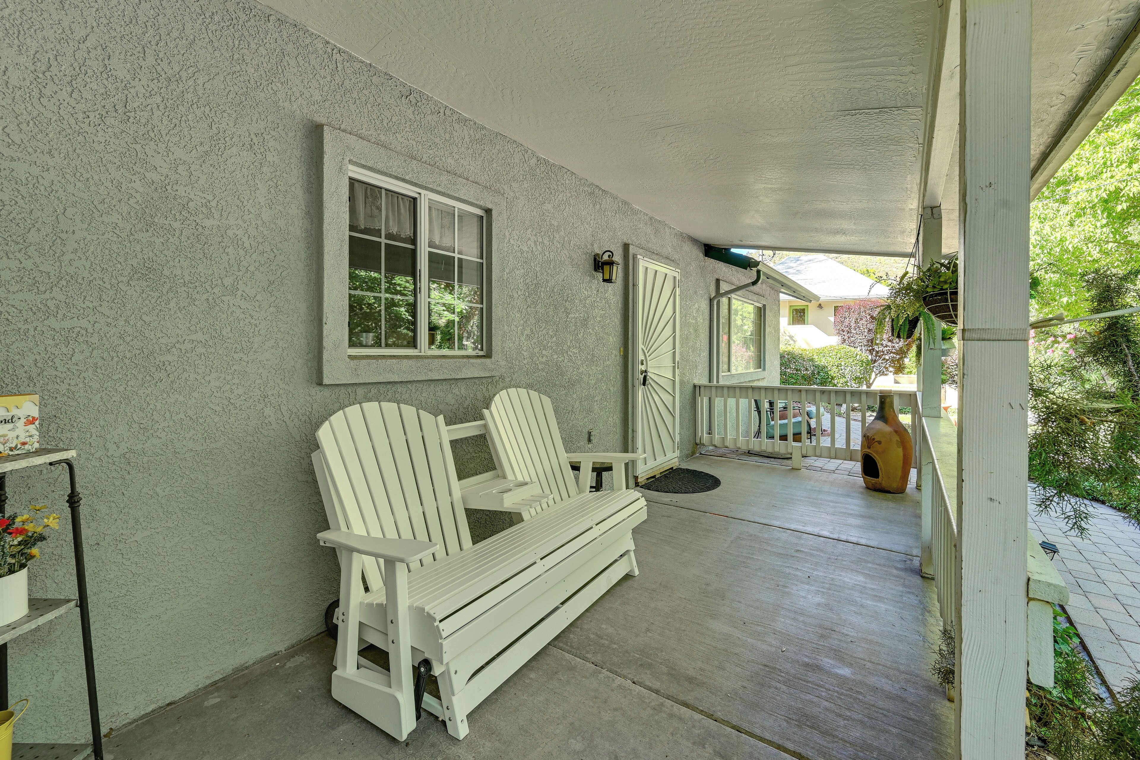 A covered porch with a white bench and Adirondack chairs, a potted plant, and a birdhouse.