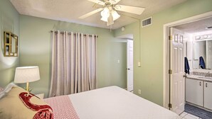 A bedroom with a bed, a ceiling fan, a bathroom with a sink and mirror, and a door leading to another room. - Cozy Beachfront Escape w/ Pool & Gulf Access! (Gulf Shores)