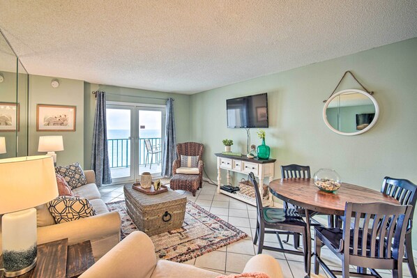 A living room with a sofa, armchair, dining table, and chairs. There is a television mounted on the wall, a window with curtains, and a mirror on the wall. - Cozy Beachfront Escape w/ Pool & Gulf Access! (Gulf Shores)