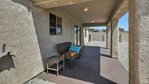 Um espaço ao ar livre coberto, com uma cadeira de vime, uma pequena mesa, piso de concreto e parede texturizada.