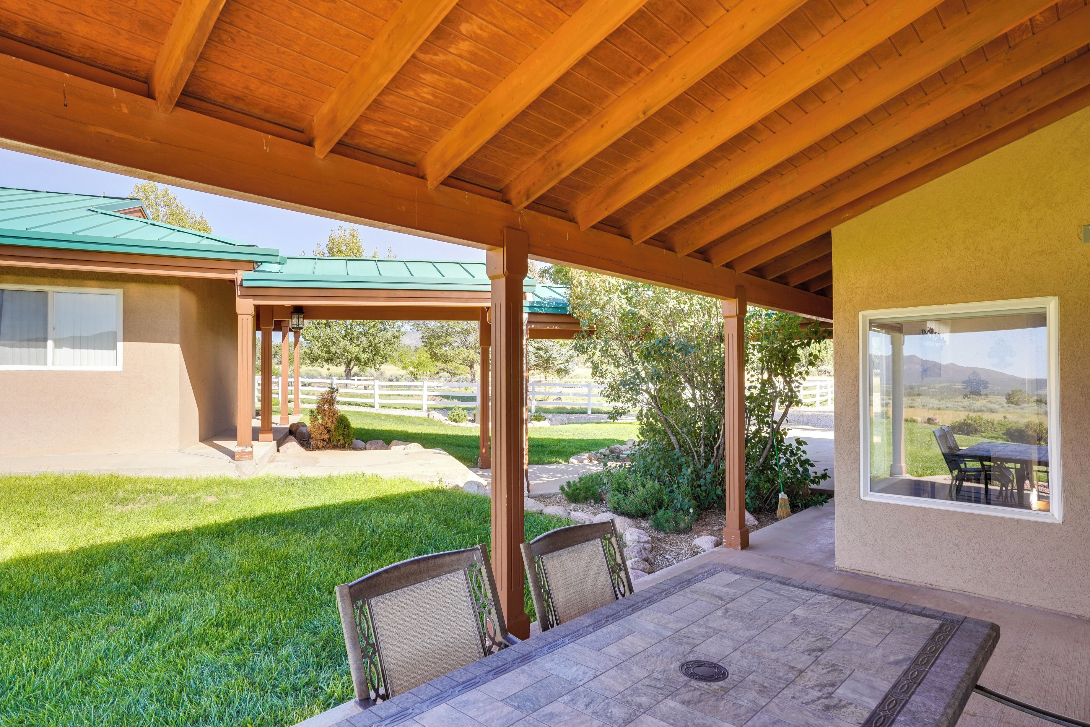 A covered patio with a table and chairs, a view of a green lawn, and a building with a green roof.