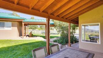 A covered patio with a table and chairs, a view of a green lawn, and a building with a green roof.