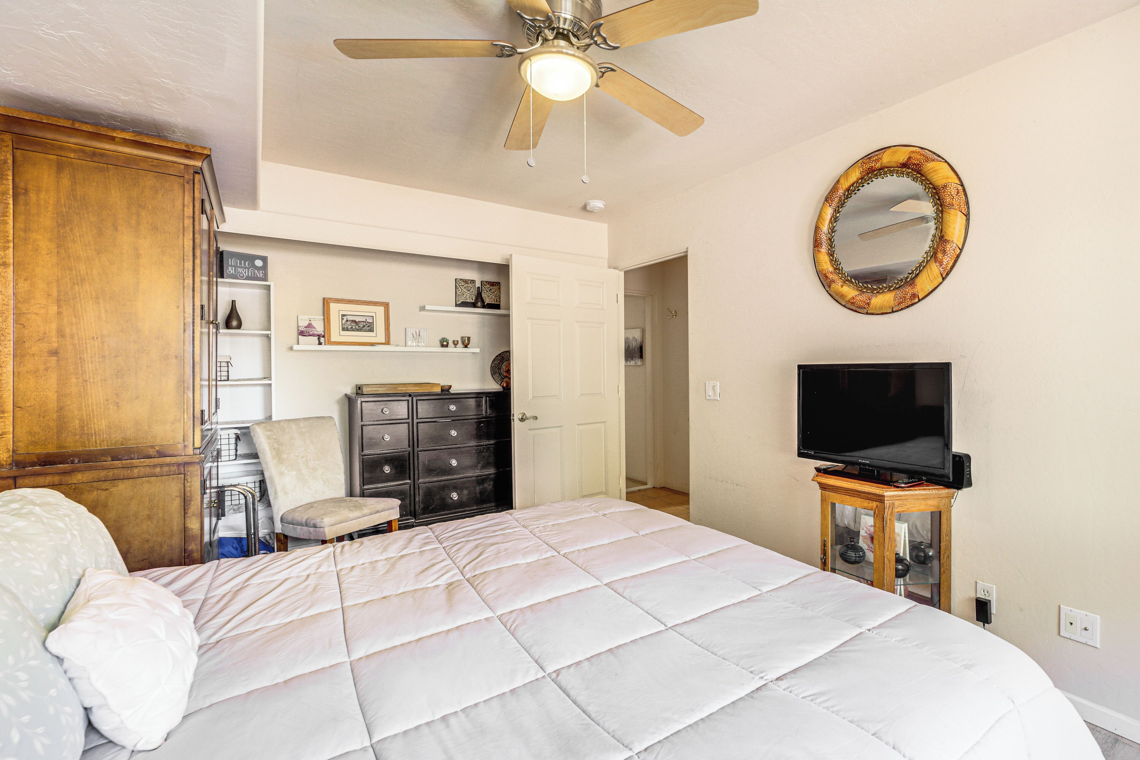 A bedroom with a large bed, a ceiling fan, a television on a wooden stand, a mirror on the wall, and a wardrobe.
