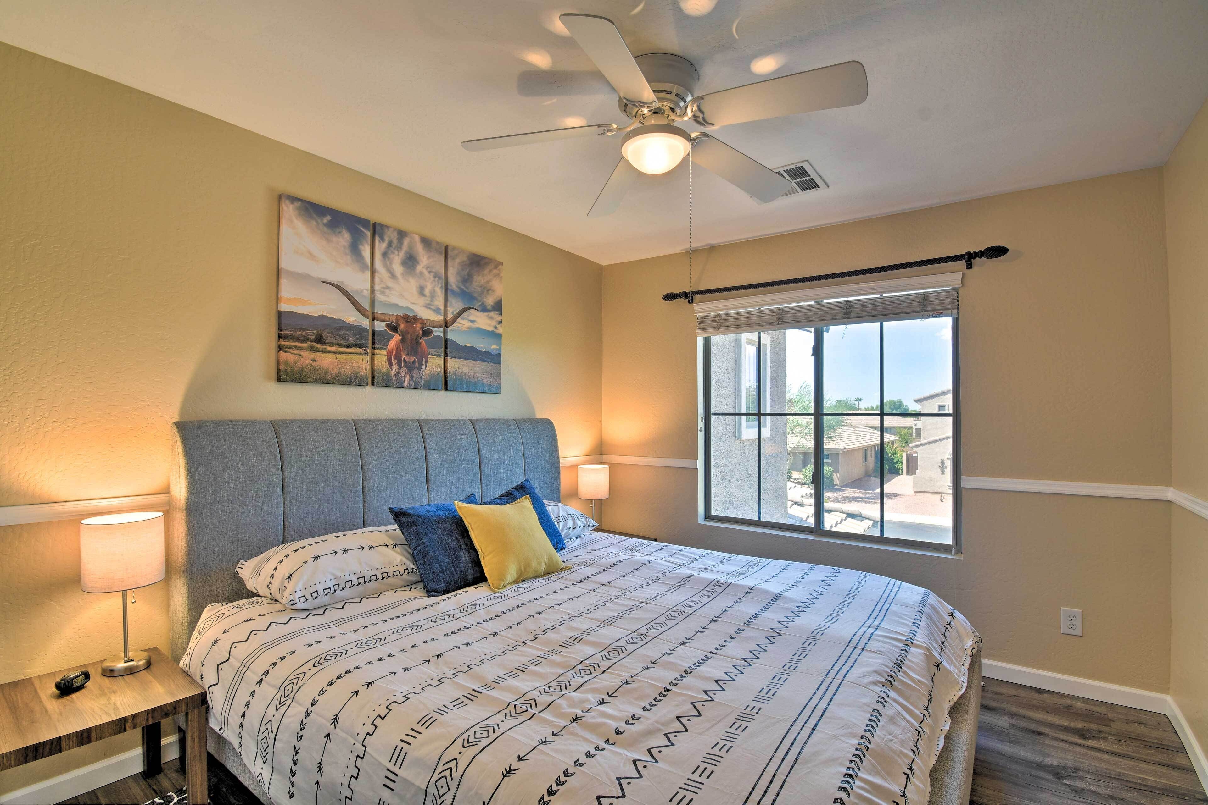 A bedroom with a bed, a ceiling fan, a large window with blinds, and a painting on the wall.