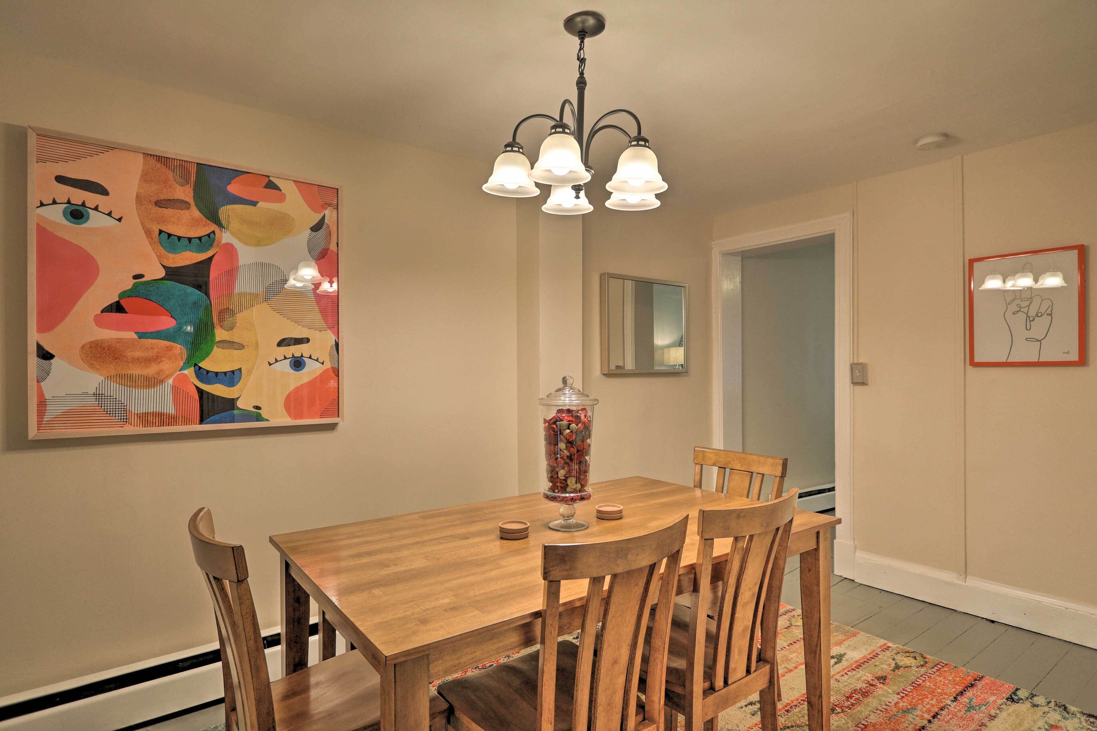 A dining area with a wooden table, four chairs, a chandelier, a large abstract painting, a mirror, and a framed picture on the wall.