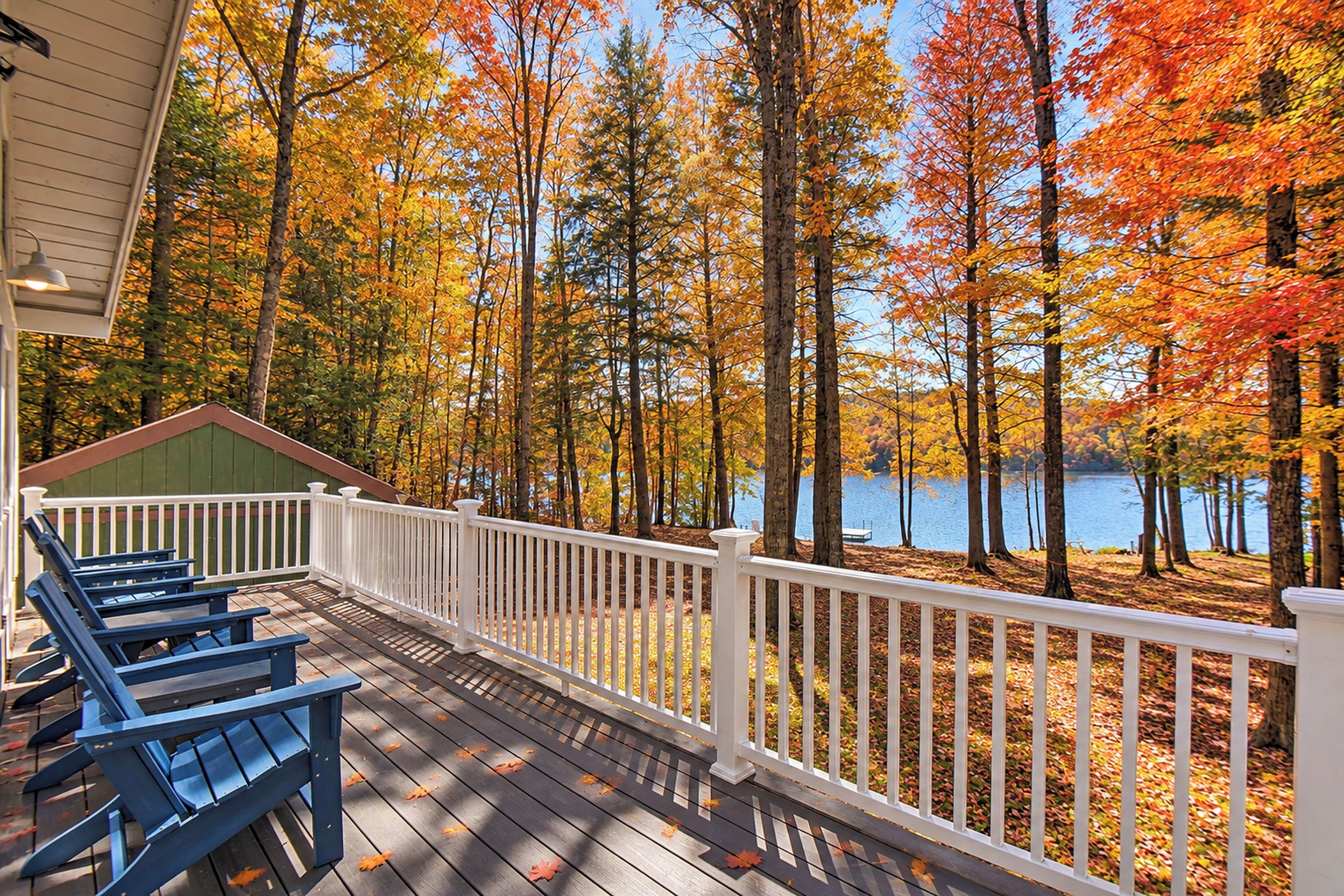 Eine Holzterrasse mit weißer Reling, die auf einen See mit herbstlich gefärbten Bäumen blickt.