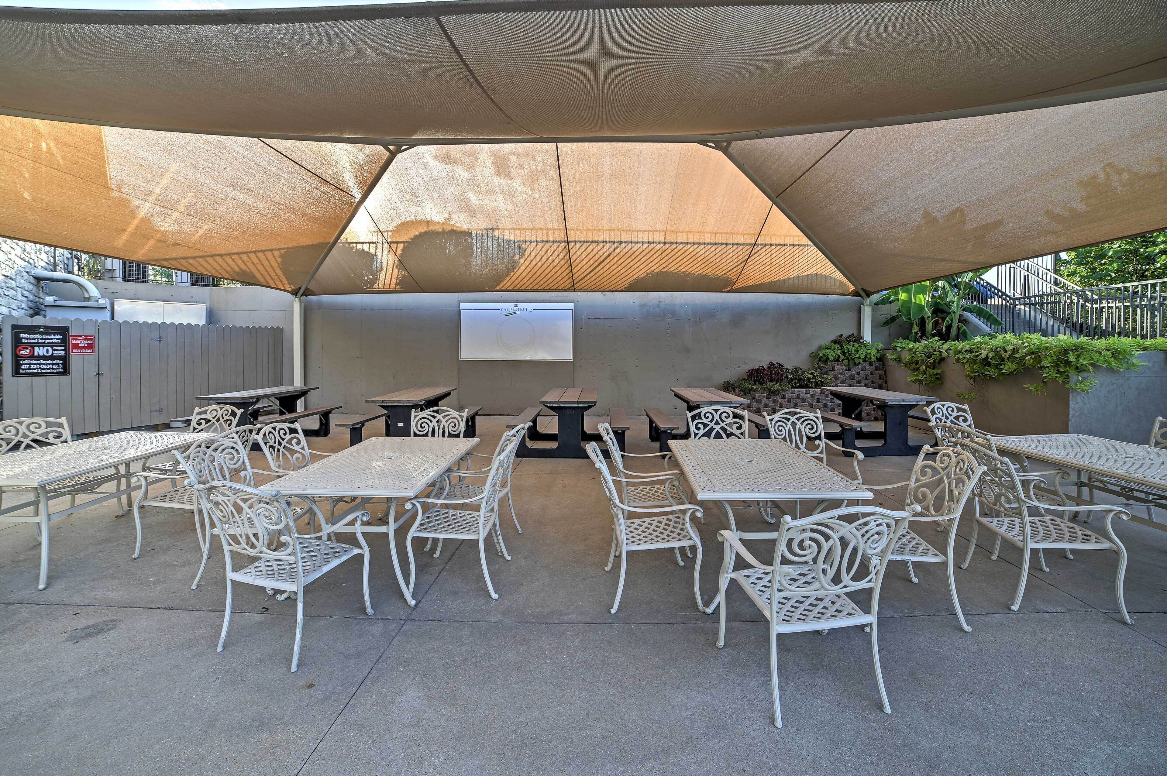 An outdoor patio area with white metal tables and chairs, a covered seating area, and a small stage with a screen.