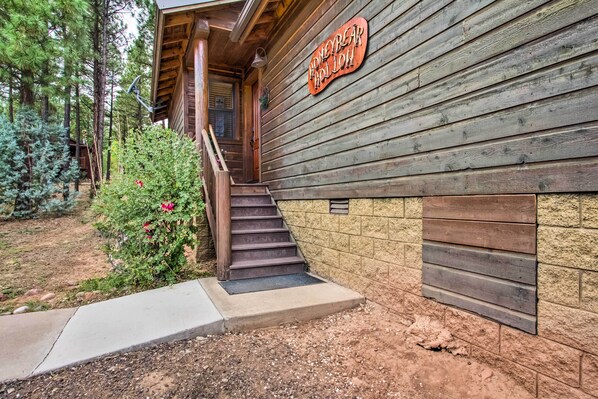 A wooden cabin with a staircase leading to a porch, surrounded by trees and a small garden with red flowers.