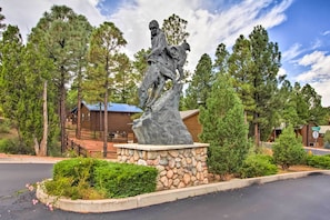 Estátua de um homem a cavalo numa área ajardinada, com pinheiros e um edifício de madeira ao fundo.