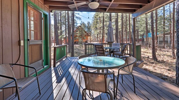 A covered outdoor deck with a table and chairs, surrounded by trees and a cabin in the background.