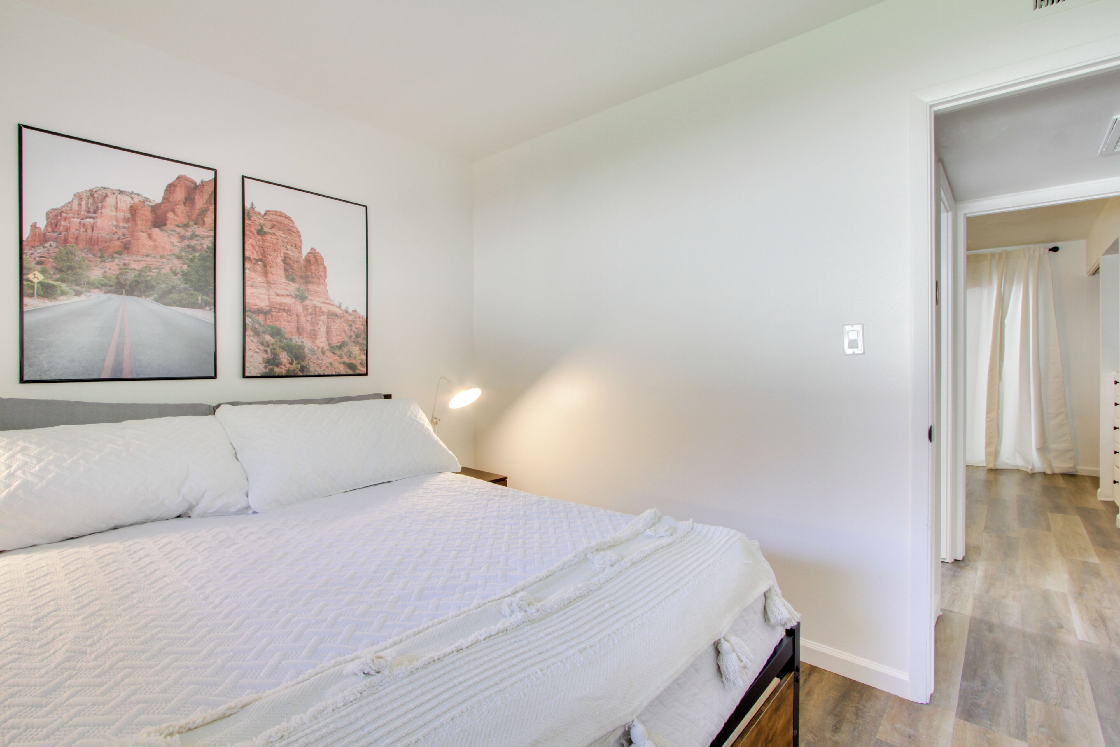 Quarto com uma cama grande, duas fotografias emolduradas na parede, uma mesa de cabeceira e uma porta que dá para outro cômodo.