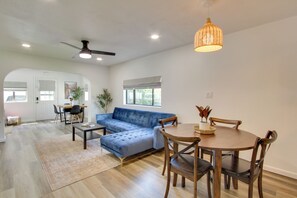 Amplia sala de estar con sofá azul, mesa de comedor de madera y sillas. Se aprecia un ventilador de techo, una lámpara colgante y un gran ventanal que proporciona luz natural.