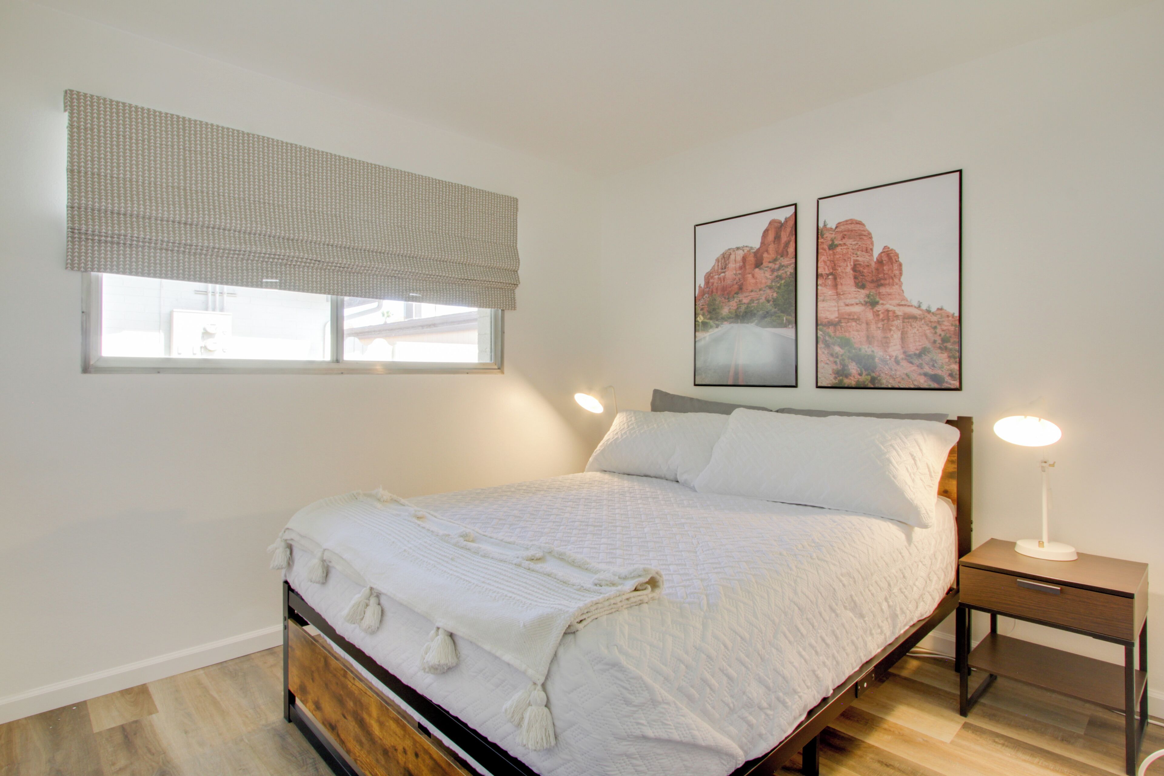 Un dormitorio con cama, mesita de noche, ventana con persiana y dos cuadros enmarcados en la pared.