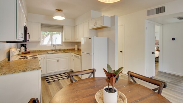 Eine moderne Küche mit weißen Schränken, einer Granitabdeckung, einem eingebauten Herd und einem Kühlschrank. Ein Esstisch mit Stühlen und einem Blumentopf steht daneben.