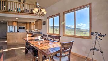 A spacious dining area with a wooden table and chairs, a kitchen in the background, and large windows offering a mountain view.