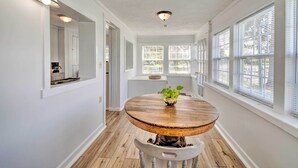 A dining area with a round wooden table, white walls, and large windows with blinds. - Romantic Waterfront Abode With Patio & Dock! (Deltaville)