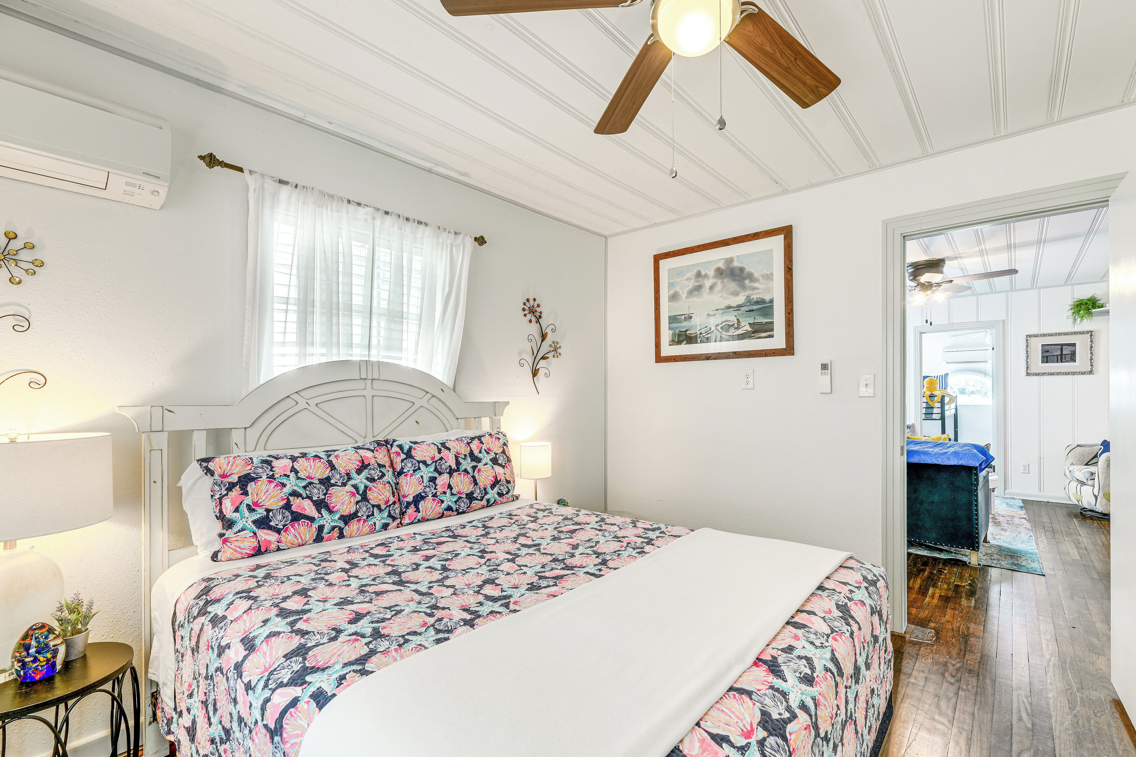 A bedroom with a bed, a ceiling fan, a window with curtains, a bedside table with a lamp, and a door leading to another room.