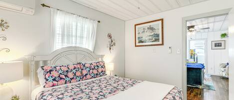 A bedroom with a bed, a ceiling fan, a window with curtains, a bedside table with a lamp, and a door leading to another room.