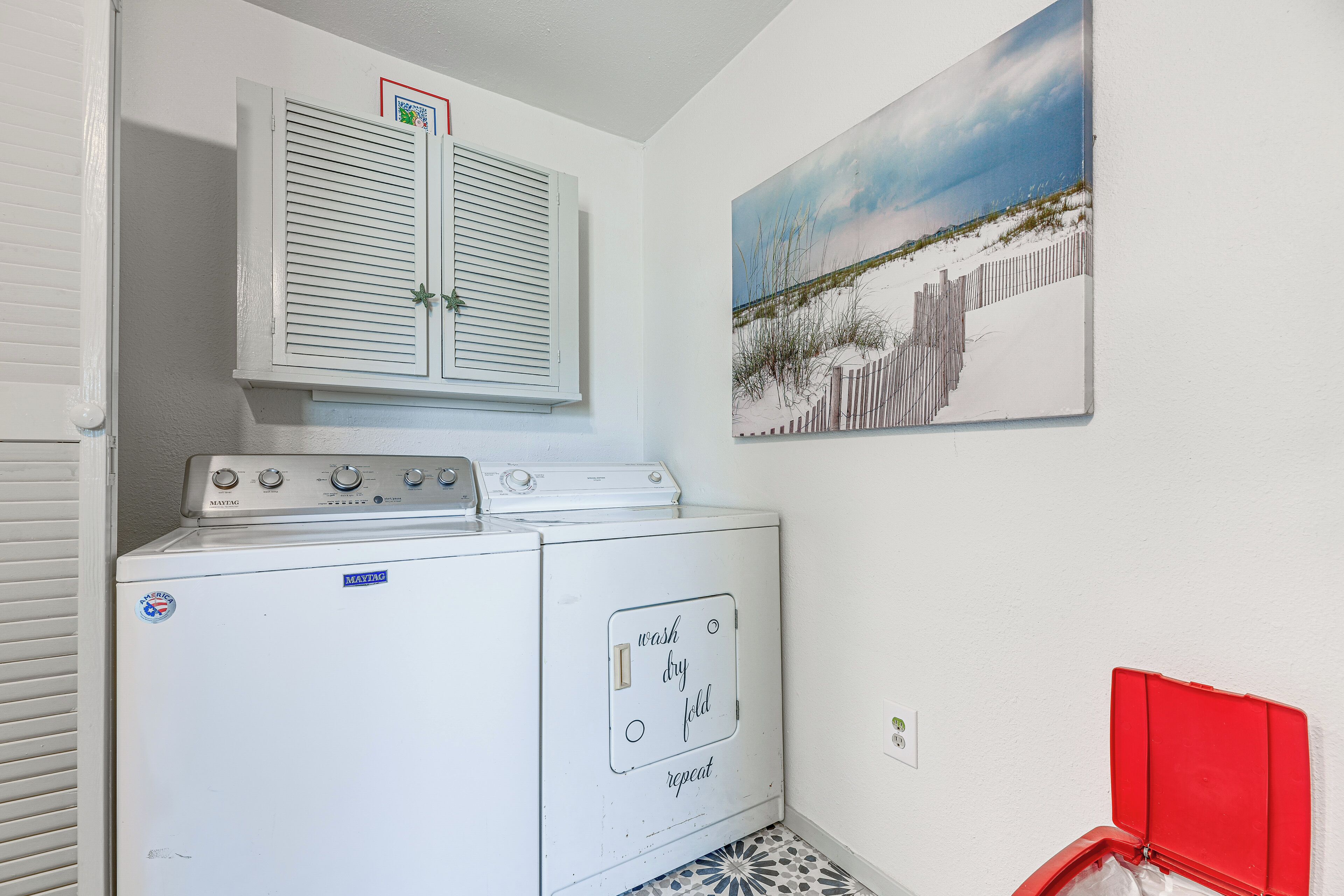 A laundry area with a front-loading washer and dryer, a window with blinds, and a beach scene painting on the wall.
